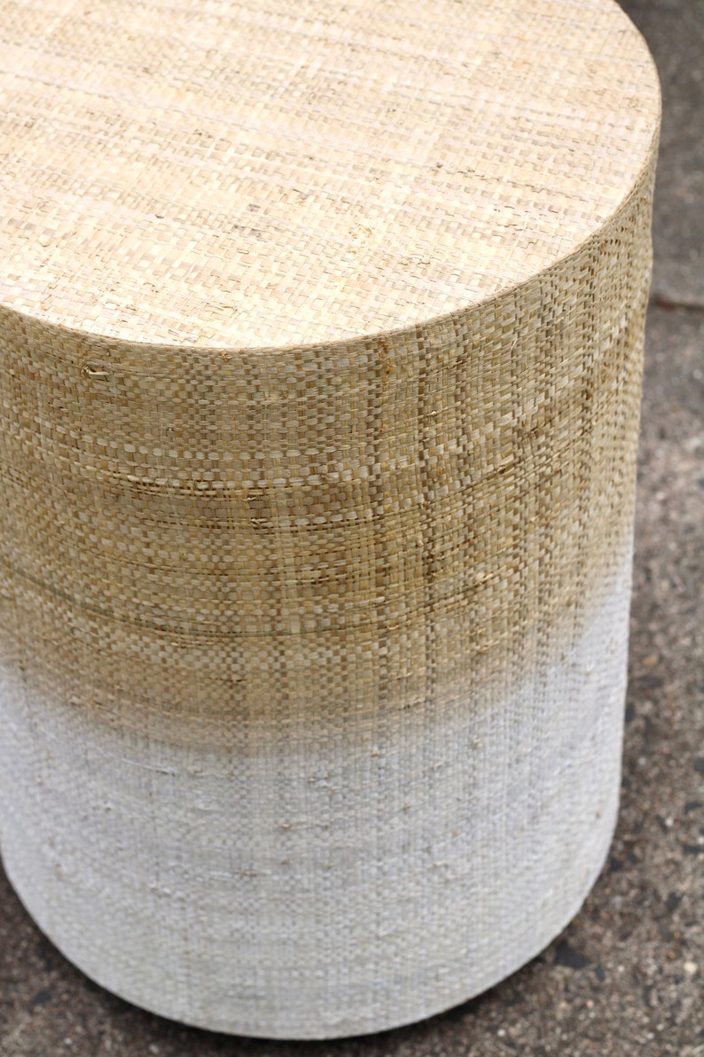 May include: A round, woven stool with a natural beige top and a white bottom. The stool is made of a woven material that gives it a rustic look.