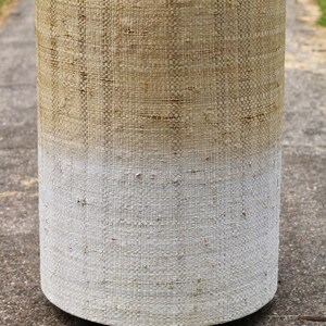 May include: A cylindrical woven stool with a light brown top that fades to white at the bottom. The stool has a woven texture and is sitting on a gray surface.