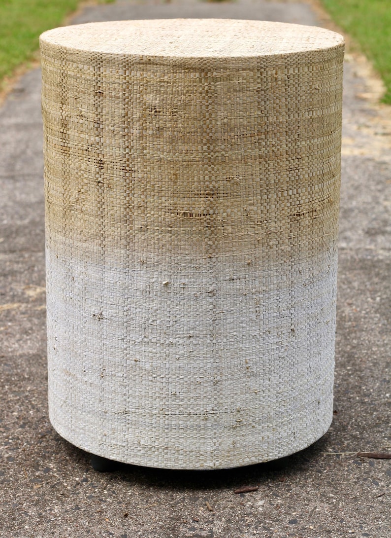 May include: A cylindrical woven stool with a light brown top that fades to white at the bottom. The stool has a woven texture and is sitting on a gray surface.