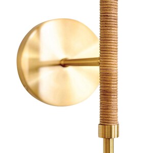 May include: A wall sconce featuring a circular, brushed brass backplate and a vertical, textured, light brown rattan-wrapped rod. The sconce has brass accents and a minimalist design, suitable for interior lighting.