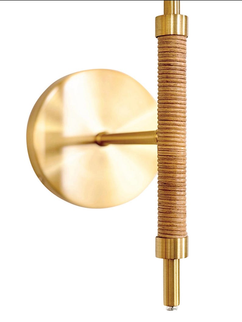 May include: A wall sconce featuring a circular, brushed brass backplate and a vertical, textured, light brown rattan-wrapped rod. The sconce has brass accents and a minimalist design, suitable for interior lighting.