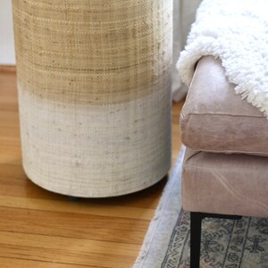 May include: A round woven stool with a light brown top and a white bottom. The stool is sitting on a wooden floor with a rug underneath.