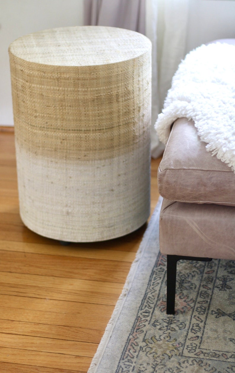 May include: A round woven stool with a light brown top and a white bottom. The stool is sitting on a wooden floor with a rug underneath.