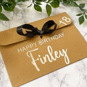 以下が含まれることがあります： 黒いリボンが結ばれた茶色の紙の封筒。封筒には「Happy Birthday Finley」と「18」の数字が印刷されています。