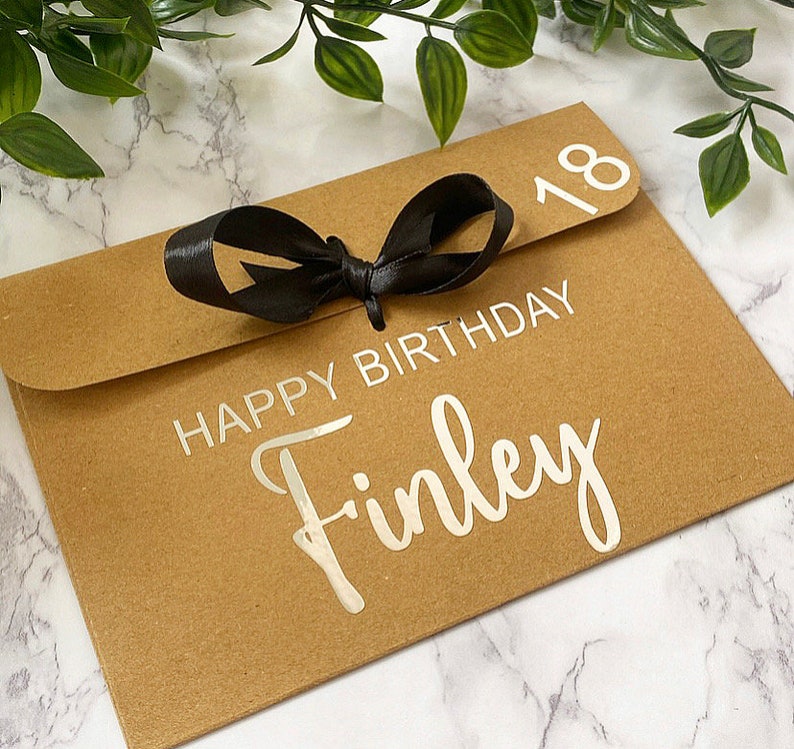 以下が含まれることがあります： 黒いリボンが結ばれた茶色の紙の封筒。封筒には「Happy Birthday Finley」と「18」の数字が印刷されています。