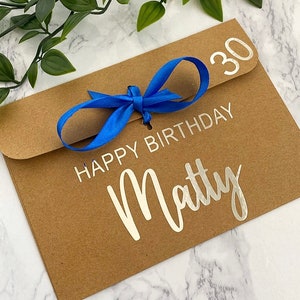 以下が含まれることがあります： 青いリボンが結ばれた茶色の紙の封筒。封筒には「Happy Birthday Matty 30」という銀色の文字が書かれています。