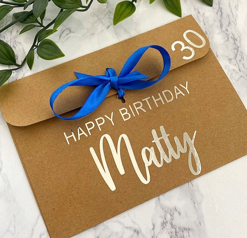 以下が含まれることがあります： 青いリボンが結ばれた茶色の紙の封筒。封筒には「Happy Birthday Matty 30」という銀色の文字が書かれています。