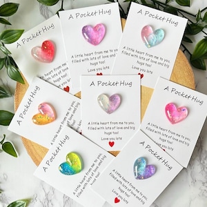 May include: Seven white rectangular cards each featuring a small, glossy, heart-shaped resin charm in various colours including pink, blue, yellow, and iridescent tones. Each card displays the text A Pocket Hug and a printed message with a red heart.