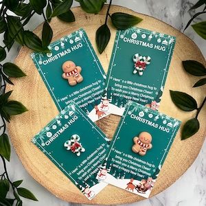 以下が含まれることがあります： 「CHRISTMAS HUG」と書かれた4枚の長方形のカード。それぞれ、赤と白のマフラーをしたジンジャーブレッドマン、または赤いリボンのついたキャンディケインの小さなフィギュアが付いています。カードにはテキストとクリスマスのイラストが印刷されています。