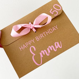 以下が含まれることがあります： ピンクのリボンと「Happy Birthday Emma 50」の文字が印刷された茶色の紙製のギフトバッグです。
