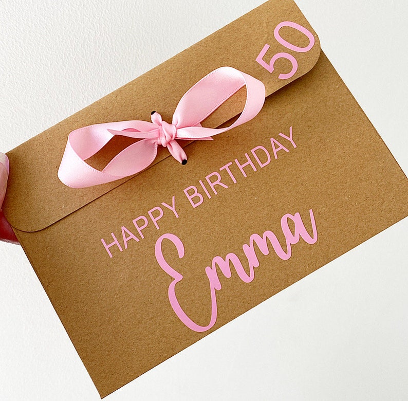 以下が含まれることがあります： ピンクのリボンと「Happy Birthday Emma 50」の文字が印刷された茶色の紙製のギフトバッグです。
