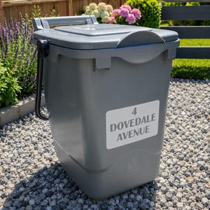 以下が含まれることがあります： グレーのプラスチック製屋外用ゴミ箱。ヒンジ付きの蓋とハンドルが付いています。「4 DOVEDALE AVENUE」と書かれたラベルが付いています。砂利の上に置かれ、背景には緑があります。