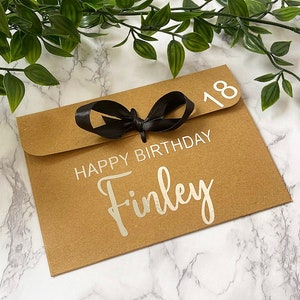 以下が含まれることがあります： 黒いリボンが結ばれた茶色の紙の封筒。封筒には「Happy Birthday Finley」という文字と「18」という数字が白で印刷されています。