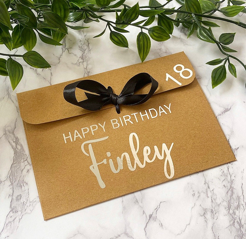 以下が含まれることがあります： 黒いリボンが結ばれた茶色の紙の封筒。封筒には「Happy Birthday Finley」という文字と「18」という数字が白で印刷されています。