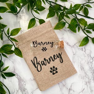 May include: A small glass bottle with a cork stopper, filled with a white substance. The bottle is labelled with the name "Barney" and a paw print. The bottle is in a hessian bag with the name "Barney" and a paw print.