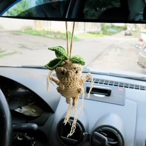 Op de afbeelding: Een gehaakte Mandragora-plant die aan de achteruitkijkspiegel van een auto hangt. De plant is beige met groene bladeren en lange, draderige wortels.