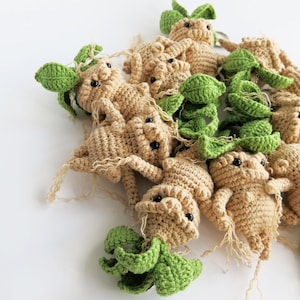 Pode incluir: Um grupo de bonecas de raiz de mandrágora de crochê com folhas verdes. As bonecas são bege e têm olhos pretos.