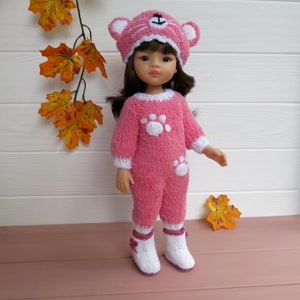 May include: A pink knitted doll outfit with white paw prints and a matching pink knitted hat with cat ears. The doll is wearing white knitted boots.