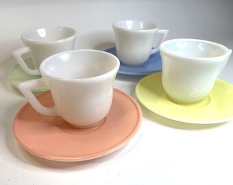 May include: Four white ceramic teacups and saucers in different pastel colors. The cups are all the same size and shape, and the saucers are all different colors. The colors are light blue, light green, light yellow, and light pink.