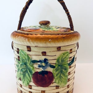 May include: A ceramic jar with a woven handle, decorated with a basket weave pattern and a painted tomato, leaves, and flower. The jar has a lid with a knob.