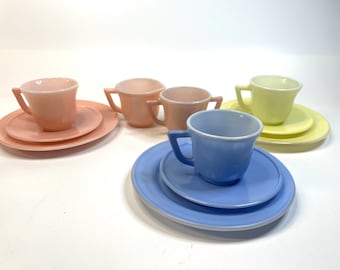 May include: A set of four pastel-colored teacups and saucers. The cups are pink, yellow, blue, and light pink. The saucers are white with a thin pastel-colored rim.