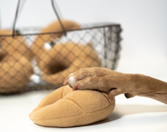 May include: A tan dog paw rests on a tan plush toy shaped like a loaf of bread. A wire basket filled with similar toys sits in the background. The toys are designed for dogs.