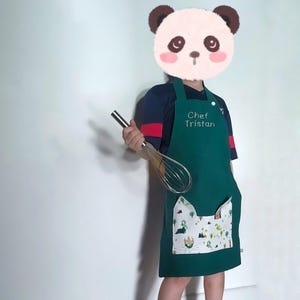 Peut inclure: Un tablier de chef vert avec une poche blanche ornée d'un motif de chat de dessin animé. Le tablier porte le texte "Chef Tristan" brodé sur le devant.