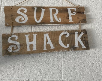 May include: A wooden sign with the words "SURF SHACK" painted in white on it. The sign is hanging from a rope and is against a white textured wall.