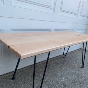 Könnte beinhalten: Ein heller Holztisch mit natürlicher Oberfläche und schwarzen Haarnadelbeinen. Der Tisch ist aus massivem Holz gefertigt und hat ein schlichtes, modernes Design.