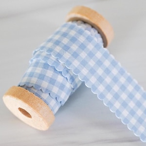 May include: A spool of light blue and white gingham ribbon with a scalloped edge. The ribbon is wrapped around a wooden spool. The ribbon is unwound, showing the pattern and texture.