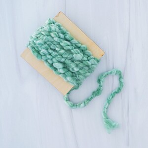 May include: A skein of light green yarn wound around a wooden spool. The yarn is soft and fluffy, and the spool is made of natural wood.