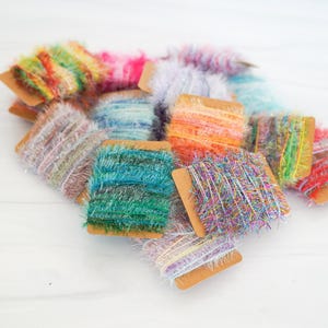 May include: A collection of colorful yarn spools. The spools are made of various colors, including green, blue, orange, pink, and purple. The yarn has a fuzzy texture and is wrapped around small cardboard spools. The spools are arranged on a white surface.