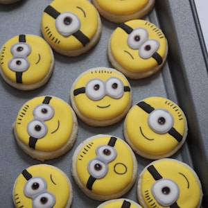 May include: A tray of yellow and black Minion-themed sugar cookies. The cookies are decorated with white eyes, black goggles, and a black mouth.