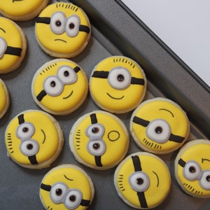 May include: A baking sheet filled with yellow and black Minion-themed cookies. The cookies are decorated with white icing and black lines to resemble the characters' eyes, mouths, and goggles.
