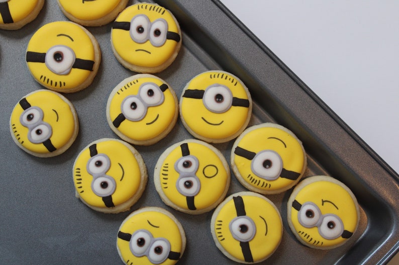 May include: A baking sheet filled with yellow and black Minion-themed cookies. The cookies are decorated with white icing and black lines to resemble the characters' eyes, mouths, and goggles.