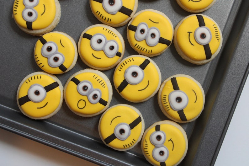 May include: A collection of yellow and black Minion-themed cookies decorated with white icing and black details. The cookies are arranged on a gray baking sheet.