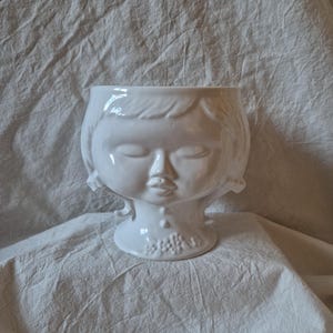 Peut inclure: Un cache-pot en céramique blanche brillante en forme de tête humaine avec les yeux fermés et une petite bouche. Les cheveux sont coiffés au carré avec des boucles sur les côtés, et la base présente un petit détail floral en relief sur le cou.