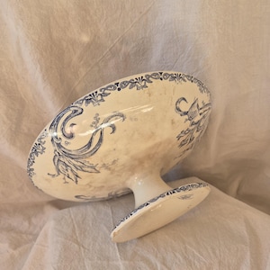 Op de afbeelding: Een crèmekleurige keramische schaal op voet met een blauw transferware-patroon van florale krulmotieven en decoratieve randen. De schaal heeft een brede, ondiepe kom die wordt ondersteund door een stevige, ronde voet.