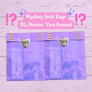 Op de afbeelding: Twee lavendelkleurige mystery grab bags met gouden hartvormige "bedankt" stickers. De tassen zijn gelabeld "Mystery Grab Bags BL/Anime/Yaoi themed". Elke tas heeft een goud-wit gestreepte sluiting.
