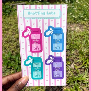 Puede incluir: Una hoja de pegatinas con cuatro botellas de lubricante de colores con el texto "Knotting Lube". Las pegatinas son de color rosa, azul, verde azulado y morado, con un dise&ntilde;o de salpicaduras blanco. El fondo es de rayas rosas y blancas con peque&ntilde;os corazones rosas.