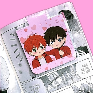 May include: A pink bookmark featuring two anime-style characters in red jackets, surrounded by heart shapes. The bookmark is placed on top of a manga page with Korean text. The characters have black and red hair.