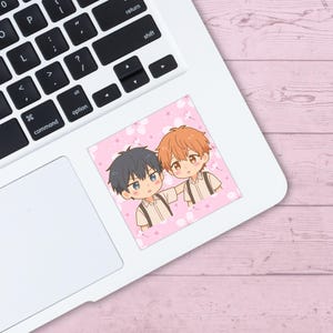 May include: A square sticker featuring two anime-style characters with dark and orange hair, set against a pink background with white floral accents. The sticker is placed on a white laptop, with a pink wooden surface in the background.