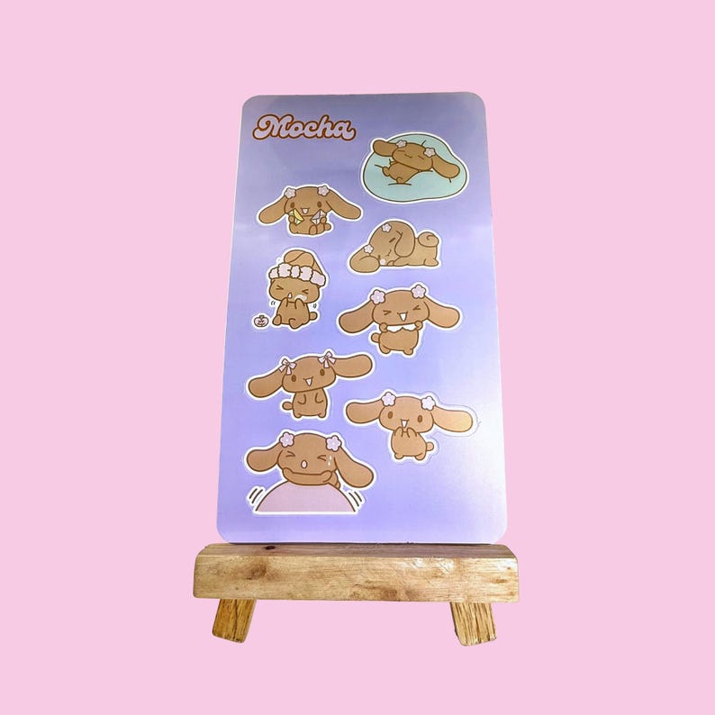 May include: A light purple sticker sheet with various cartoon dog illustrations. The word "Mocha" is written in cursive at the top. The sticker sheet is displayed on a small wooden easel made of wood.