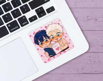 May include: A square sticker featuring an anime-style illustration of two male characters reading a book together. The book has a heart and Japanese text. The background is pink with heart shapes. The sticker is on a white laptop.