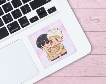 May include: A square sticker featuring two anime-style characters embracing. The characters have black and blonde hair, wearing tan shirts. The background is a pastel pink and blue with heart shapes.