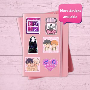 May include: A pink notebook adorned with various stickers. The stickers showcase anime-style illustrations and text, including a jar with hearts, a No-Face character, and a design with the text "M-Preg Enjoyer". A pink speech bubble says "More designs available".