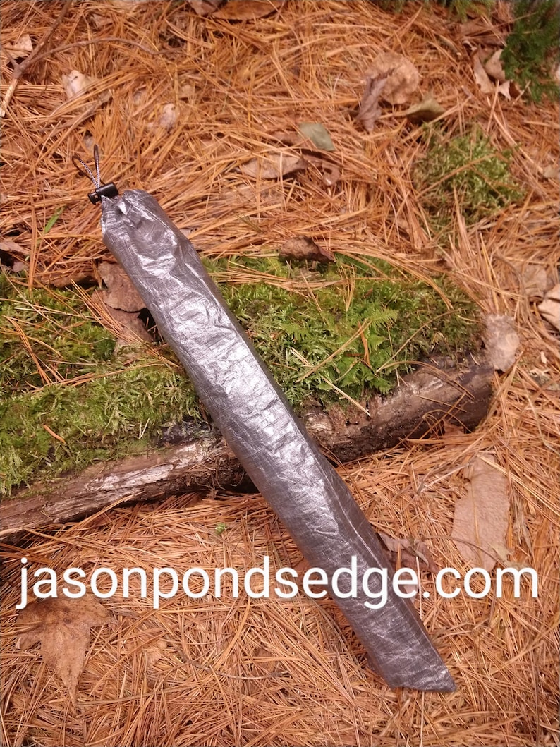 Puede incluir: Una funda rectangular plateada con cierre de cord&oacute;n, sobre un lecho de agujas de pino y musgo. El texto "jasonpondsedge.com" est&aacute; impreso en blanco en la parte inferior de la imagen. La funda parece estar hecha de un material duradero e impermeable.