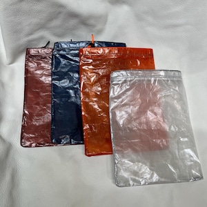 May include: Four colorful, lightweight pouches in shades of red, navy blue, orange, and silver. Each pouch has a drawstring closure at the top. The pouches are made of a translucent, crinkled material.