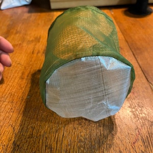 May include: A cylindrical storage bag featuring a translucent olive green ripstop mesh upper section and a solid, opaque silver-colored reinforced base. The bag has a structured, circular bottom and a flexible, lightweight construction.