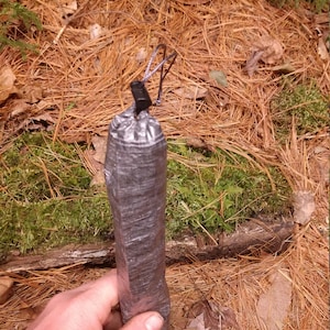 Puede incluir: Un objeto rectangular gris oscuro envuelto en cinta adhesiva, con un cierre de cord&oacute;n negro. El objeto se sostiene sobre un fondo de agujas de pino, hojas y musgo.